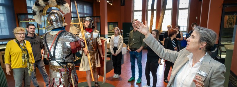 Eine Gruppe Besucherinnen und Besucher steht in der Ausstellung vom Vindonissa Museum (in der Mitte lebensgrosse Legionäre). Eine Museumsführerin steht davor und zeigt auf die beiden Männer.