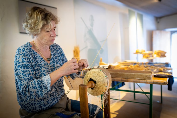 Eine Frau arbeitet mit Stroh, im Hintergrund ein Tisch mit Kunstwerken aus Stroh.