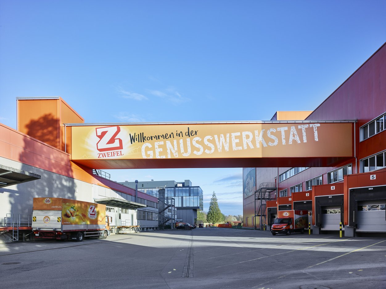 Fotografie vom Neubau in Spreitenbach. Auf dieser ist die vorwiegend in Orange gehaltene Aussenanlage mit den orangen Zweifel-Transportern zu sehen. 