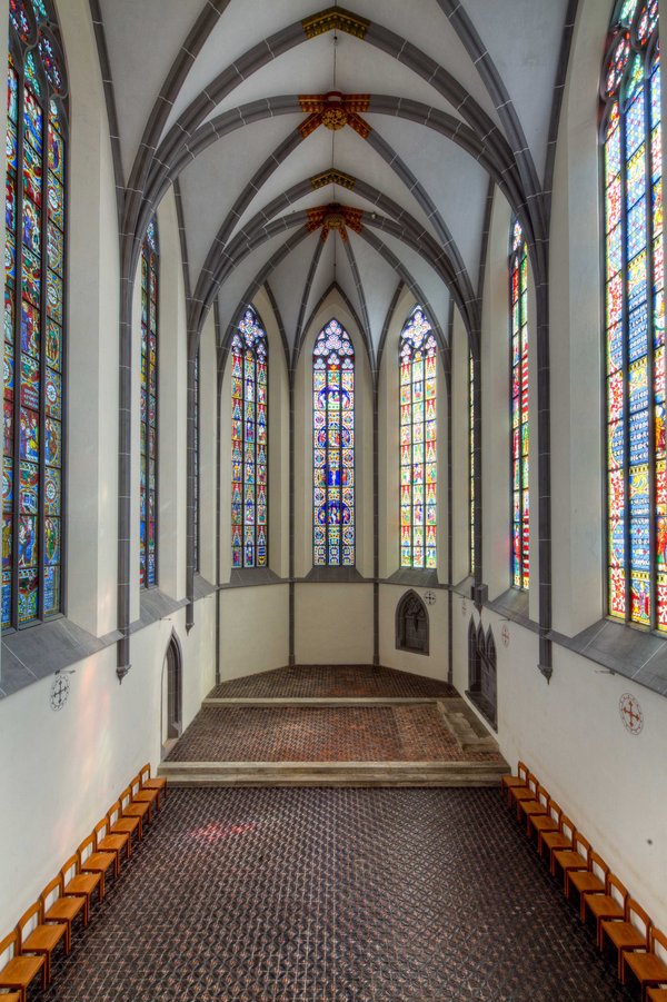 Das Bild zeigt einen Kirchenchor mit farbenfrohen Glasmalereien an den Fenstern und einer gewölbten Decke. 