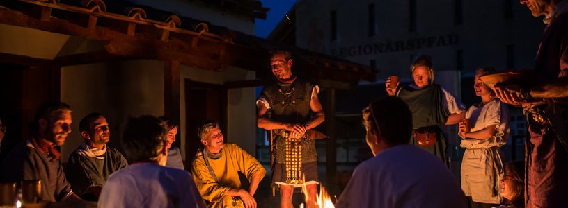 Römisch gekleidete Männer und Frauen nachts an einem Lagerfeuer in historischer Kulisse