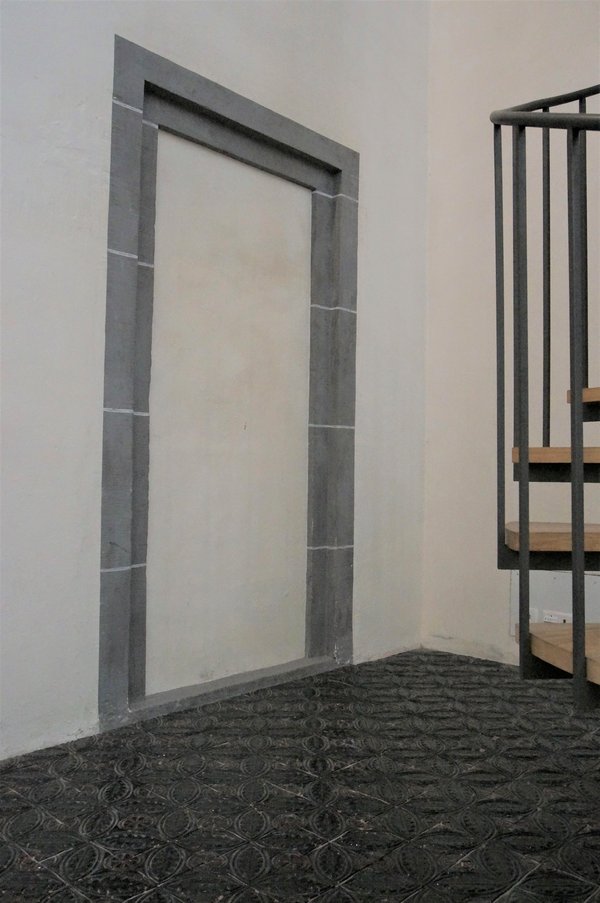 Das Bild zeigt einen Eingang mit einem schwarzen Boden. Auf der rechten Seite des Bildes ist ein Teil einer Wendeltreppe erkennbar. 