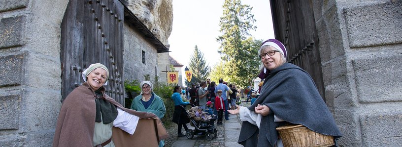 Zwei historisch gekleidete Freuen stehen vor dem Tor von Schloss Lenzburg und bitten Besucher mit einer einladenden Geste herein.
