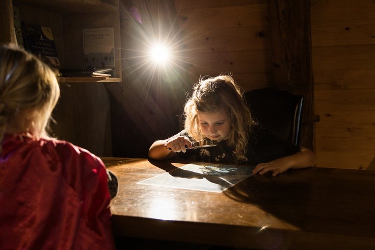 Ein Mädchen untersucht im Kindermuseum von Schloss Lenzburg mit einer Lupe ein Dokument; von hinten erleuchtet ein Scheinwerfer die Szenerie im mystischen Gegenlicht. 