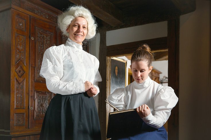 Zwei historisch gekleidete Damen in einem Raum von Schloss Lenzburg. Eine blickt direkt in die Kamera, die andere ist versunken in einem Buch.