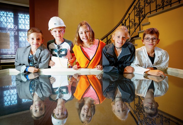 Kinder als Forscher und Forscherinnen gekleidet lehnen sich an einen spiegelnden Tisch