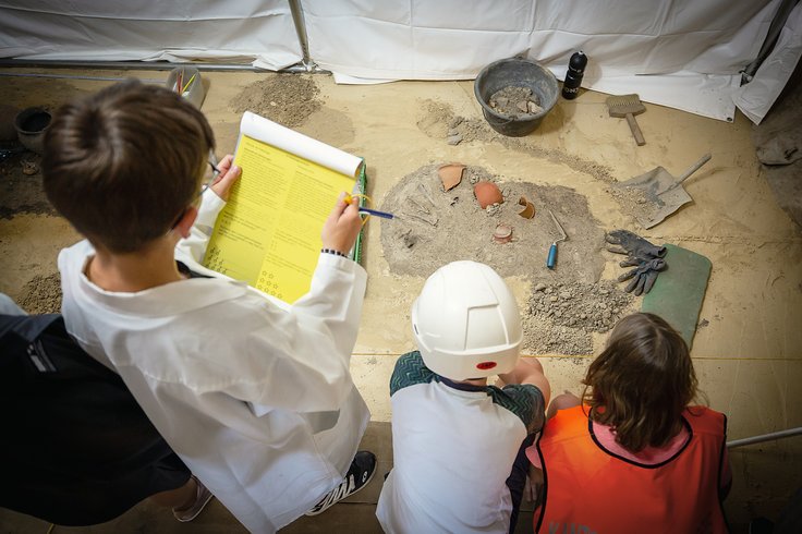 Kinder als Forscher*innen verkleidet begutachten die Ausstellung und lösen Rätsel (Aufnahme von Oben)