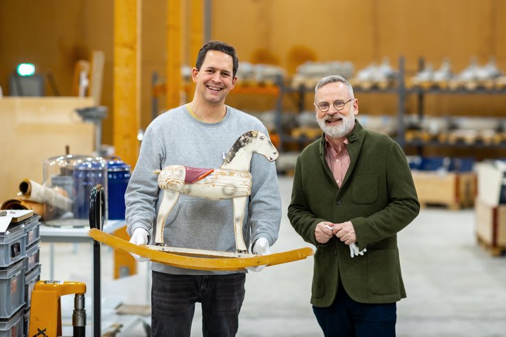 Zwei Männer stehen in einer Halle im Sammlungszentrum Egliswil und blicken lächeln in die Kamera. Der linke Mann hält ein grosses Schaukelpferd aus Holz in der Hand.