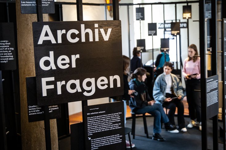 Blick in den Ausstellungsraum "Parlatorium" auf der Klosterhalbinsel Wettingen. Im Vordergrund ein Schild mit dem Text "Archiv der Fragen", im Hintergrund sitzen Schülerinnen und Schüler diskutierend beeinander.