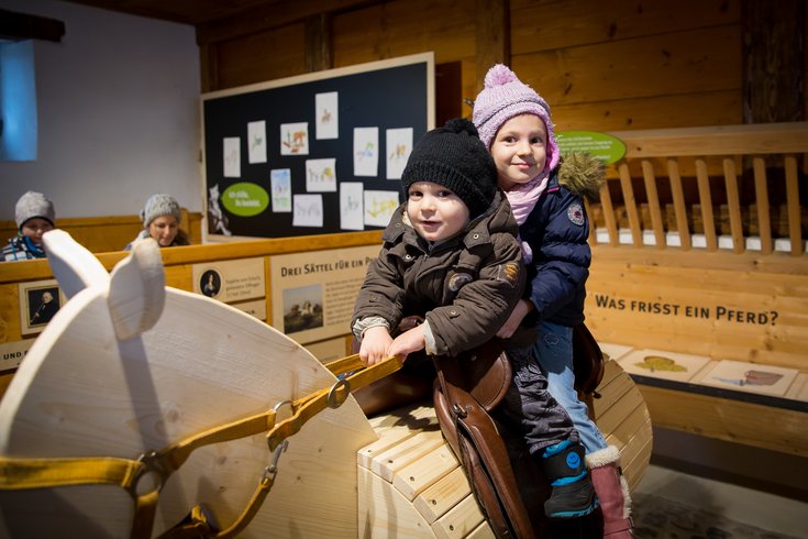 Mädchen und Junge reiten auf einem Holzpferd im Pferdestall auf Schloss Wildegg