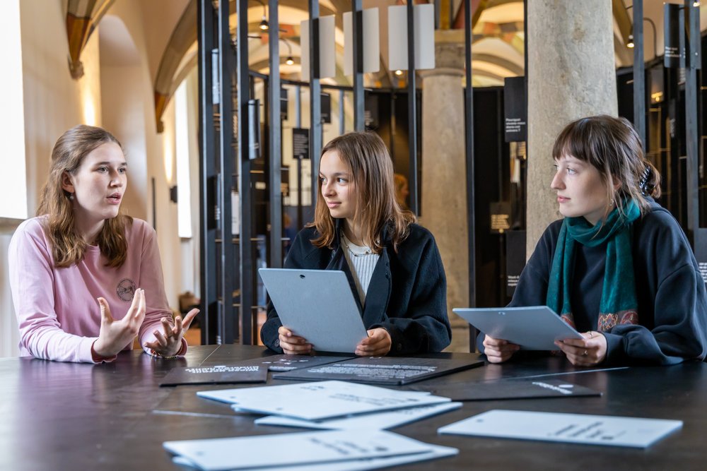 Ausstellung "Parlatorium auf der Klosterhalbinsel Wettingen": Drei Schülerinnen sitzen an einem Tisch und diskutieren angeregt über Fragen, die auf grossen Tafeln vor ihnen stehen.
