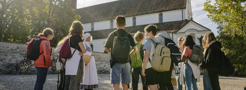 Eine Klostermagd steht mit einer Gruppe Schülerinnen und Schülern vor dem Kloster Wettingen.