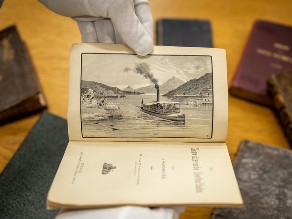 Ein Paar weisse Handschuhe hält ein geöffnetes Buch aus der Schlossbibliothek Wildegg. Auf der Seite ist die Abbildung eines Dampfschiffs zu sehen.