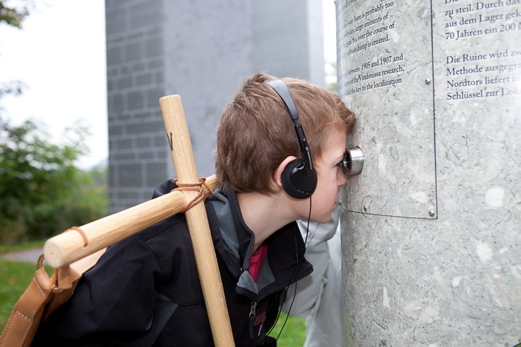 Junge mit kopfhörern und einem Stock in der Hand schaut in ein Guckloch 