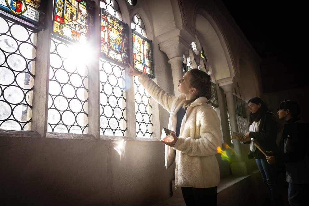 Eine Schülerin im Kreuzgang des Klosters Wettingen. Sie steht vor den Glasfenstern, durch die das Gegenlicht der Sonne in den Kreuzgang fällt.