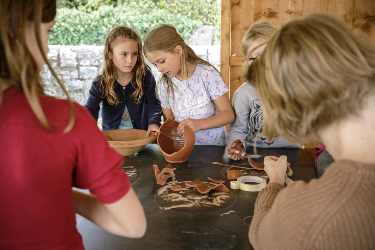 Kinder setzen an einem Tisch Scherben eines Tongefässes wieder zusammen.