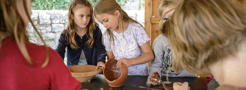 Kinder setzen an einem Tisch Scherben eines Tongefässes wieder zusammen.