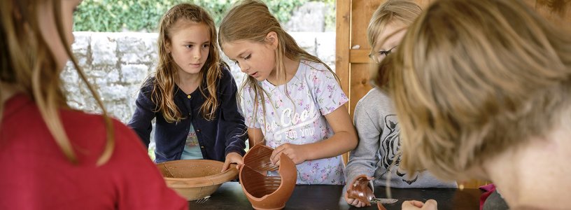 Kinder setzen an einem Tisch Scherben eines Tongefässes wieder zusammen.