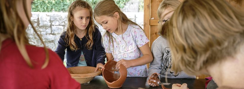 Kinder setzen an einem Tisch Scherben eines Tongefässes wieder zusammen.