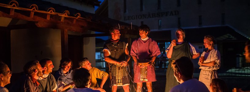 Besucher und Besucherinnen umkreisen gemeinsam mit den Legionären ein Lagerfeuer, welcher licht und wärme für die Nacht spendet.