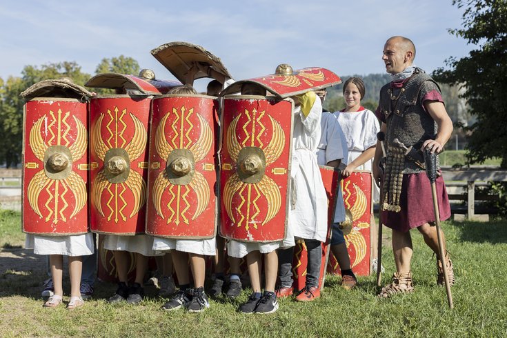 Eine Schulklasse in römischer Formation Schildkröte, rechts davon gibt ein Legionär Anweisungen.