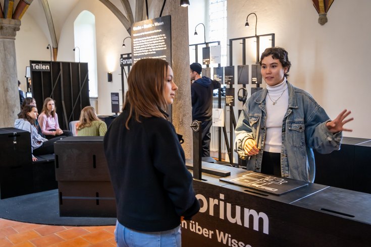 Ausstellung "Parlatorium auf der Klosterhalbinsel Wettingen": Zwei Schülerinnen diskutieren in einem grossen Raum; dahinter weitere Personen sitzend.