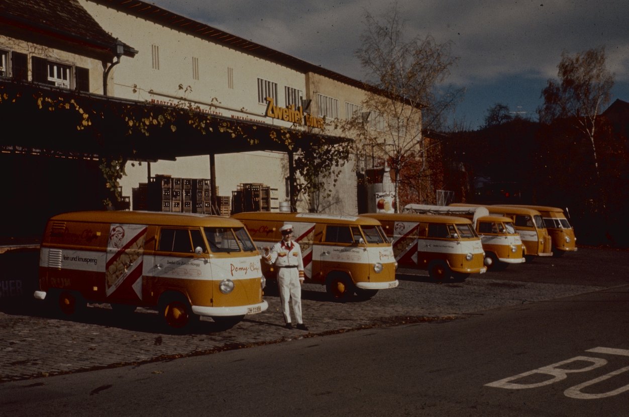 Fotografie von den damaligen VW-Bussen des Frisch-Services, welche in den Farben Orange und Weiss gehalten sind. Zudem ist auf dem Bild eine Person in der Frisch-Service-Uniform zu sehen.