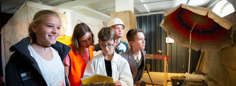 Kinder als Forscher*innen verkleidet lösen gemeinsam ein Rätsel während ein Junge im Kittel einen Klemmbrett hält