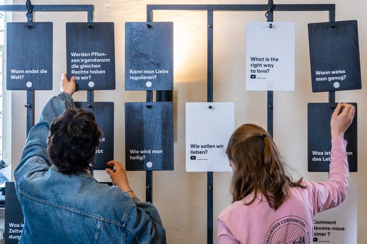 Ausstellung "Parlatorium auf der Klosterhalbinsel Wettingen": Zwei Schülerinnen greifen nach grossen Tafeln mit Fragen, die an einer Wand aufgehängt sind.