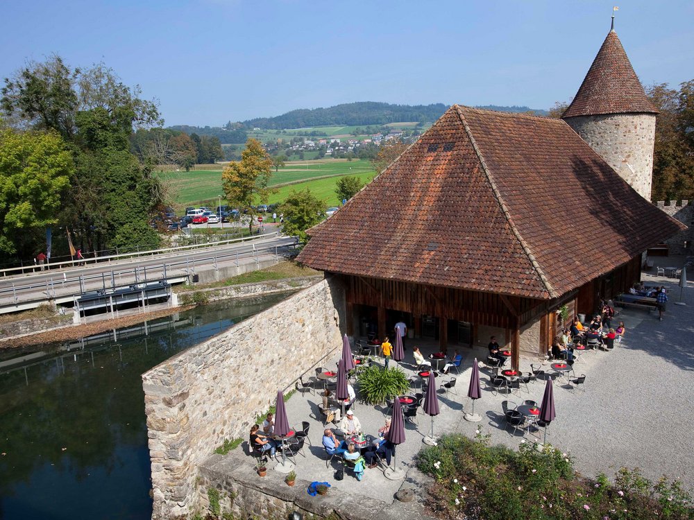 Blick von oben auf den Aussenbereich des Cafés von Schloss Hallwyl. An den Tischen im Schlosshof sitzen Besucherinnen und Besucher. Das Wetter ist sommerlich.