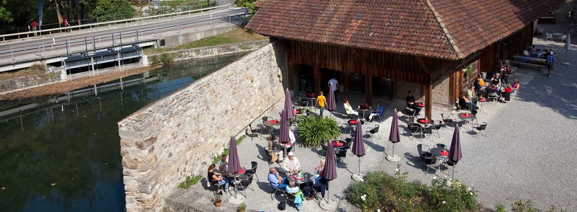 Blick von oben auf den Aussenbereich des Cafés von Schloss Hallwyl. An den Tischen im Schlosshof sitzen Besucherinnen und Besucher. Das Wetter ist sommerlich.