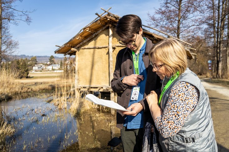 Ein Mann und eine Frau mit Foxtrail-Anhängern studieren einen Plan. Im Hintergrund das Pfahlbauhaus am Hallwilersee in Seengen.