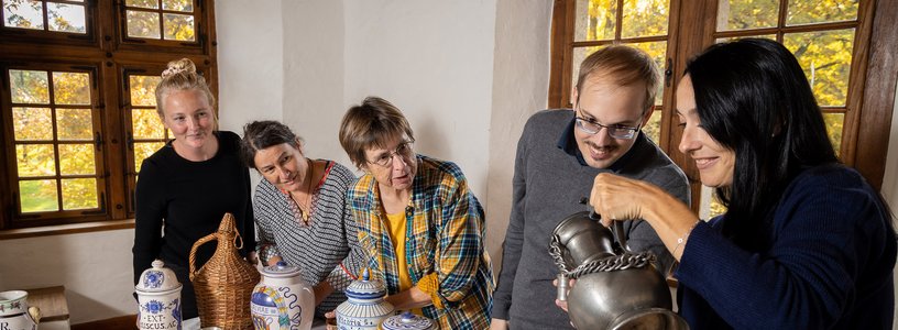 Eine Besuchergruppe in einem historischen Raum von Schloss Hallwyl. Die Gruppe steht um einen Tisch herum, auf dem Gefässe stehen. Eine Frau geisst eine Flüssigkeit aus einem Metallkrug in einen Metallbecher.