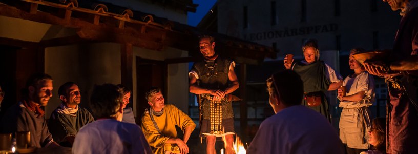 Römisch gekleidete Männer und Frauen nachts an einem Lagerfeuer in historischer Kulisse