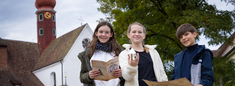 Zwei Mädchen und ein Bub stehen gemeinsam vor dem Kloster Wettingen und blicken direkt in die Kamera. Sie halten eine Landkarte, Broschüre sowie eine Pyramide in den Händen.