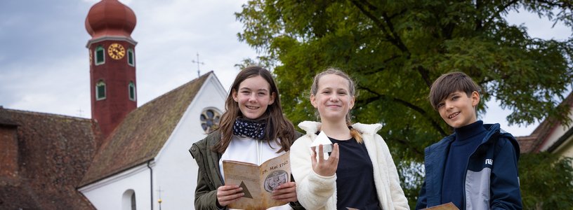 Zwei Mädchen und ein Bub stehen gemeinsam vor dem Kloster Wettingen und blicken direkt in die Kamera. Sie halten eine Landkarte, Broschüre sowie eine Pyramide in den Händen.