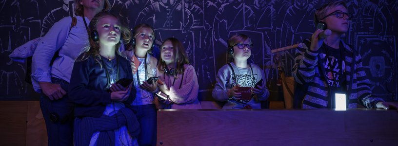 Eine Familie in einem dunklen Raum; die Displays der Audiogeräte um den Hals leuchten mystisch im Dunkeln. An der Rückwand Silhouetten von Legionären.