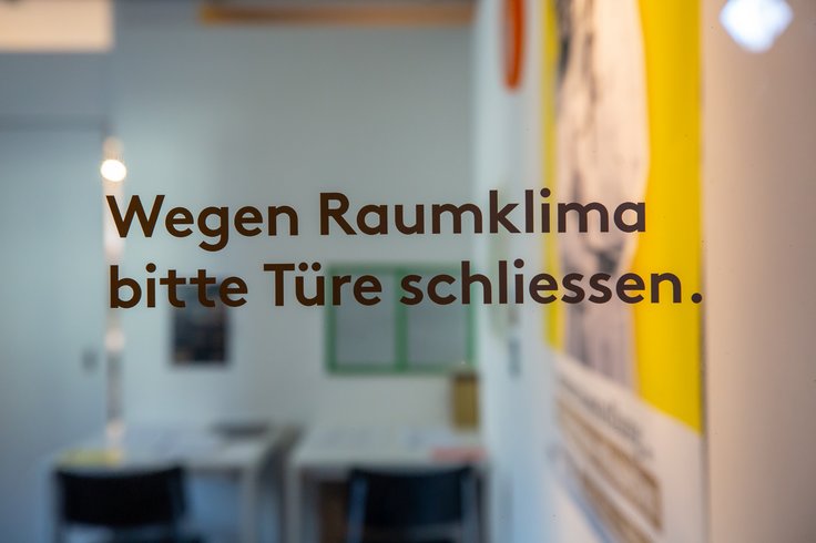 Glastüre mit der Beschriftung "wegen Raumklima bitte Türe schliessen"