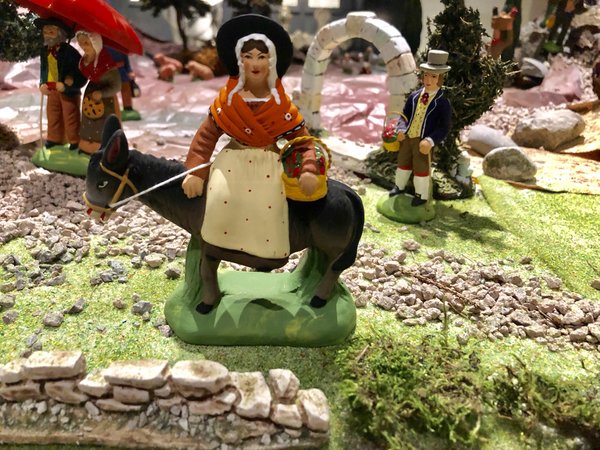 Zu sehen ist eine Nahaufnahme einer handbemalten Santons-Figur. Die Figur zeigte eine Frau, welche auf einem Esel reitet und einen Korb unter dem Arm trägt.