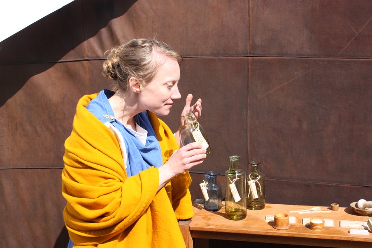Aufnahme einer Person, die an einem Glasfläschchen riecht. Auf einem Tisch im Hintergrund stehen weitere Behältnisse und Utensilien.