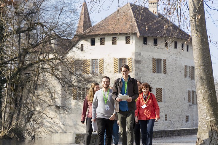 Mehrere Personen mit grünen "Foxtrail"-Anhängern, Plan und weiteren Unterlagen unterwegs auf dem Foxtrail vor Schloss Hallwyl.