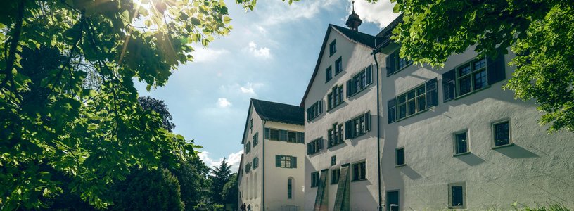 Aussenaufnahme Kloster Wettingen mit blauen Himmel, Sonne im Gegenlicht und einer grünen Wiese im Vordergrund.