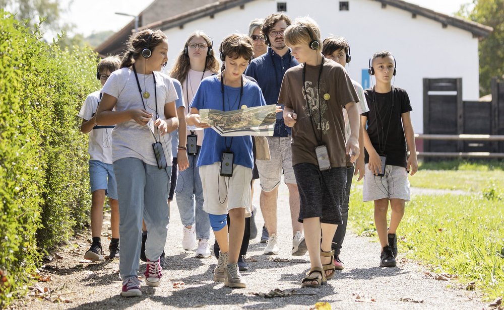 Eine Schulklasse unterwegs auf einem Kiesweg; im Hintergrund die rekonstruierte Unterkunft von Legionären.