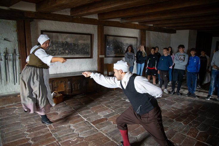 Eine Magd und ein Knecht gemeinsam mit einer Schulklasse im Schloss Wildegg. Sie zeigen einen Kampf mit Degen.