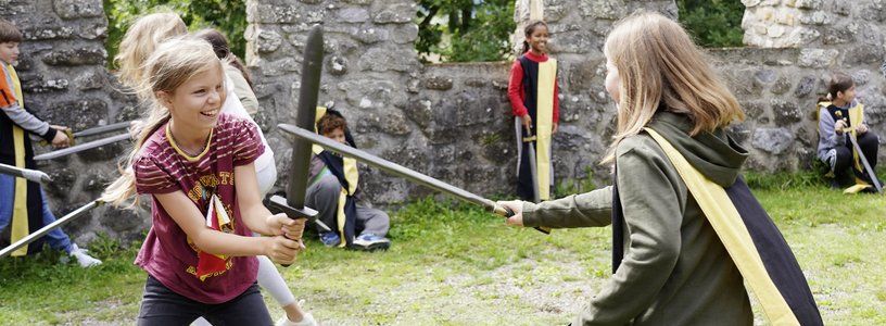 Workshop "Feste Burg Schloss Hallwyl" Kinder haben zusammen spass im Schwertkampf mit Zinnen im Hintergrund
