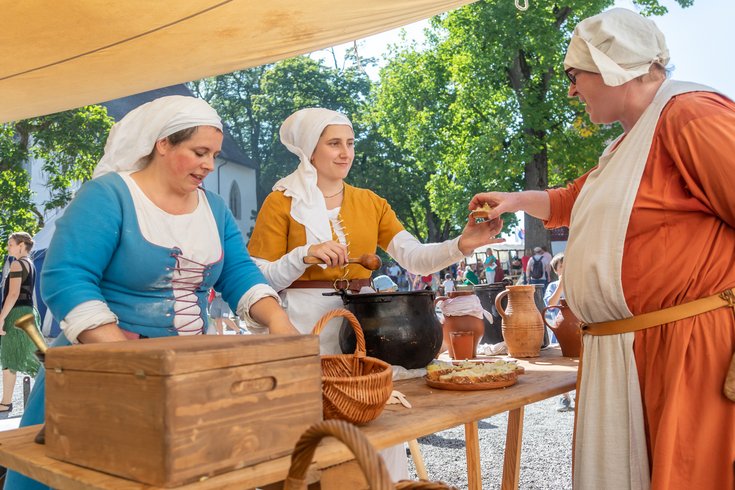 Zwei mittelalterlich gekleidete Frauen; eine übergibt einem Marktbesucher eine Speise zum Probieren.